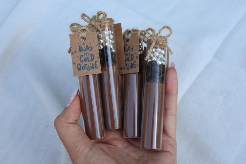 May include: A hand holds four glass tubes filled with a brown substance, each capped with silver beads and tied with twine. Small brown tags read "Baby it's Cold Outside". The tubes are a winter-themed gift.