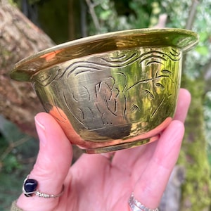 May include: A small, polished brass bowl featuring a wide, flat rim and a rounded base. The exterior surface of the bowl is decorated with hand-etched, linear patterns that wrap around the circumference, creating a textured, metallic appearance.