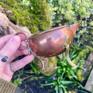 May include: A copper-colored, small, antique gravy boat with a handle and three legs. The gravy boat has a spout and is made of copper with brass accents. The handle and legs are brass-colored.
