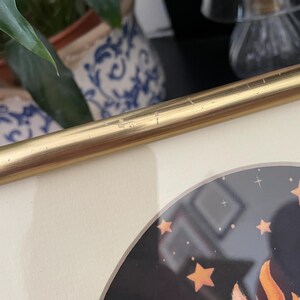 May include: A gold-toned metal picture frame with visible surface scratches along the top edge. The frame contains a circular print featuring a dark background with orange stars and a partial orange graphic element visible in the lower corner.