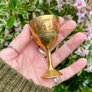 May include: A small brass goblet featuring a polished gold-tone finish with intricate etched patterns around the bowl. The design includes recessed red enamel detailing within the engravings, a slender stem, and a flared circular base for stability.