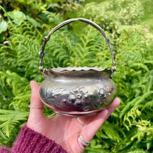 May include: A small, round, silver-toned metal basket with a scalloped rim and a swing handle. The exterior surface features an embossed floral pattern with detailed petals and leaves. The metal has a polished finish with visible signs of age.