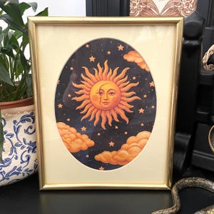 May include: A rectangular gold-toned metal frame containing an oval-shaped print. The artwork features a central orange sun with a human face, surrounded by a dark navy blue night sky filled with small yellow stars and fluffy orange clouds.