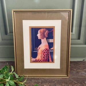 Op de afbeelding: Ingelijste prent van een Renaissance-portret. Het kunstwerk toont een vrouw in profiel, gekleed in een oranje en rode jurk met ingewikkelde details. De prent is geplaatst in een goudkleurige lijst en een witte passe-partout.