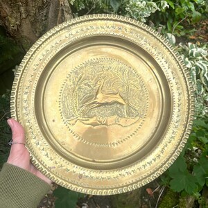 May include: A round brass decorative plate featuring an embossed central scene of a leaping deer flanked by two palm trees. The plate has a wide, textured border with a repeating geometric pattern along the outer edge and a smooth inner rim.