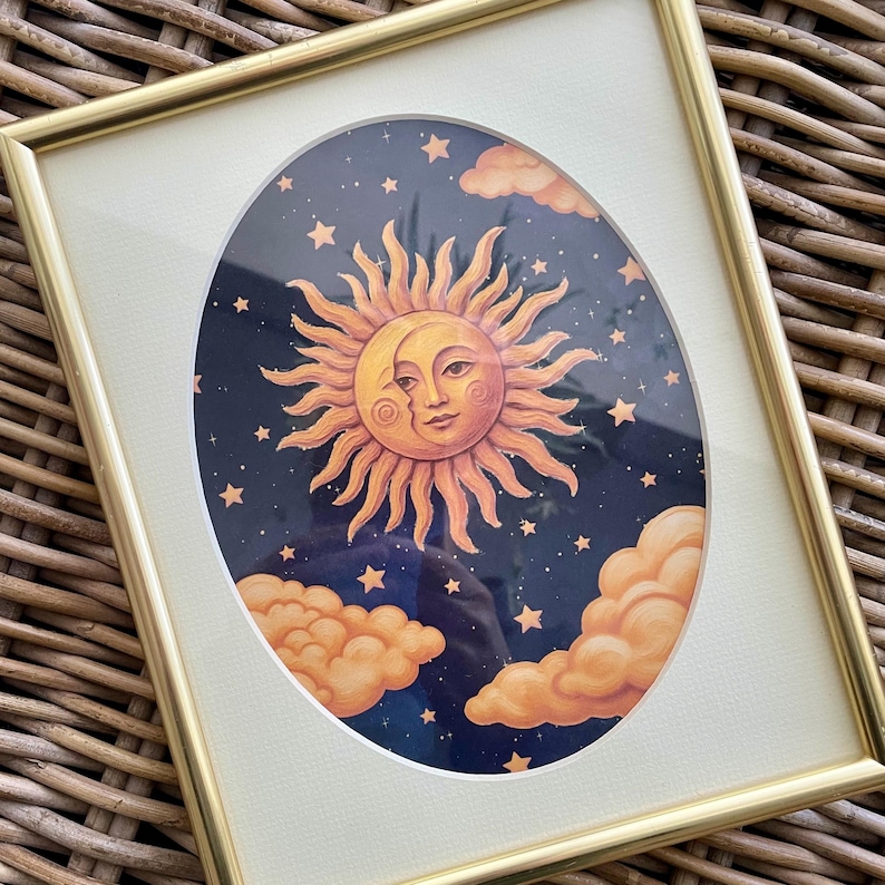 May include: A framed print featuring a central sun and moon face illustration set against a dark blue night sky filled with small stars and orange clouds. The artwork is enclosed within a thin, metallic gold rectangular frame with a white mat.