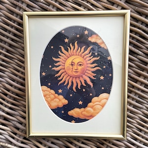 May include: A rectangular gold-toned frame containing an oval-shaped print of a sun and moon face. The illustration features a warm orange sun with a crescent moon profile against a dark blue starry night sky with orange clouds at the bottom.