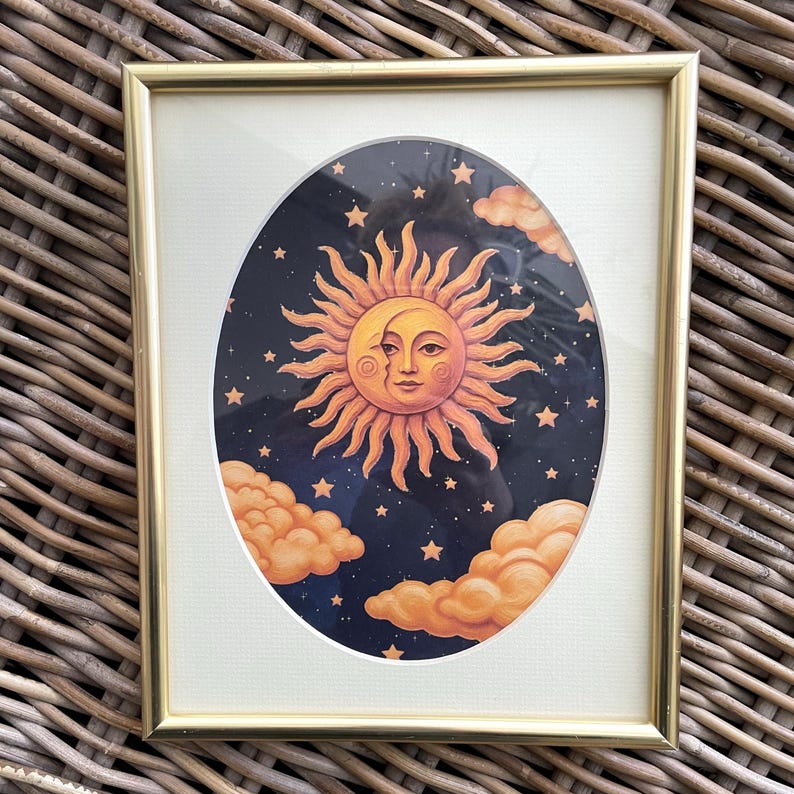 May include: A rectangular gold-toned frame containing an oval-shaped print of a sun and moon face. The illustration features a warm orange sun with a crescent moon profile against a dark blue starry night sky with orange clouds at the bottom.