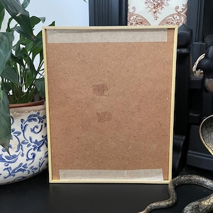 May include: A rectangular picture frame with a light-colored wooden border, shown from the back. The backing is made of brown fiberboard, secured with two horizontal strips of light-colored adhesive tape at the top and bottom edges.