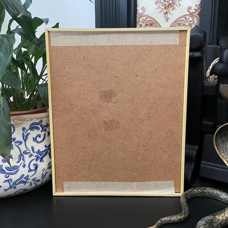 May include: A rectangular picture frame with a light-colored wooden border, shown from the back. The backing is made of brown fiberboard, secured with two horizontal strips of light-colored adhesive tape at the top and bottom edges.