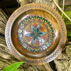May include: A round wooden plate featuring a hand-carved floral pattern in the centre with green, blue, and red painted accents. The rim is decorated with a repeating circular motif, and the wood grain is visible throughout the entire surface.