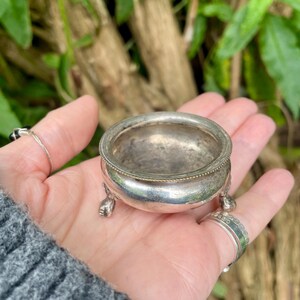 May include: A small, antique silver bowl with three decorative feet. The bowl has a rounded shape with a detailed rim. The bowl is held in a hand, with a blurred green background.