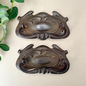 May include: Two matching dark bronze-toned metal drawer pulls featuring an ornate, symmetrical design with a central oval plate, curved decorative edges, and a hinged drop handle attached to each piece with two visible screw holes for mounting.