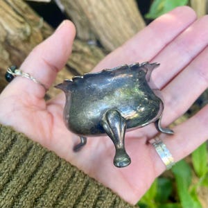 May include: A small, antique silver-colored bowl with three delicate feet and a scalloped rim. The bowl has a rounded shape and appears to be made of metal. The bowl is held in a hand, with a ring visible on the finger.
