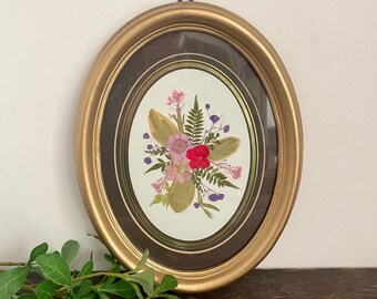 May include: Oval gold-framed pressed flower art. The artwork features a bouquet of dried flowers in shades of pink, red, and purple, with green leaves. The frame has a dark brown matting and a gold finish. A black hook is attached to the top for hanging.