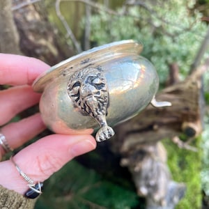 May include: A small, silver-colored metal bowl with a detailed face design on the side. The bowl has a rounded shape with a wide rim and a small foot. The face has intricate details, and the bowl appears to be vintage or antique.