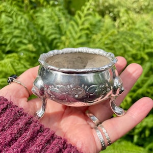 May include: A small, round, silver-toned metal bowl featuring an embossed floral pattern on its exterior. The bowl has a scalloped rim and is supported by three ornate, curved legs that terminate in detailed, sculpted animal-like feet.