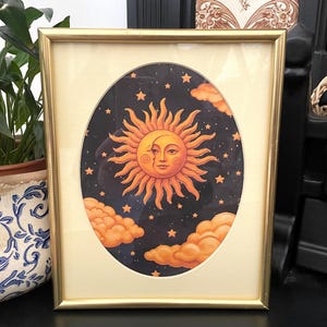 May include: A rectangular gold-toned frame containing an oval-shaped print of a sun and crescent moon face set against a dark blue starry night sky. The illustration features orange clouds at the bottom and a cream-colored mat border.