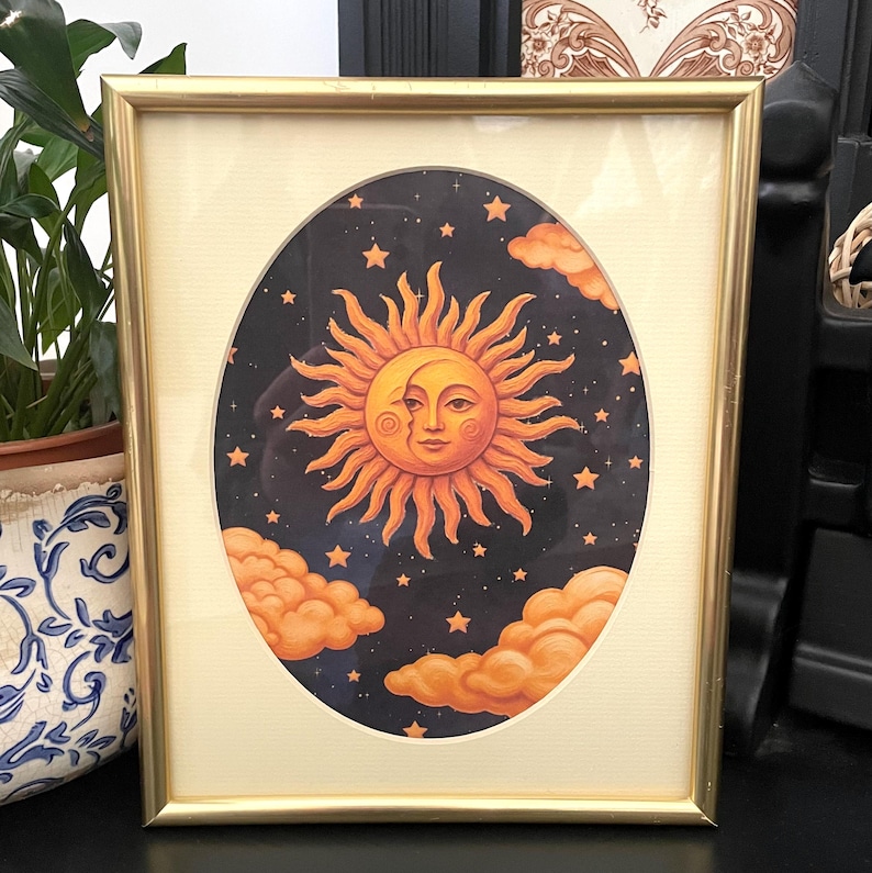 May include: A rectangular gold-toned frame containing an oval-shaped print of a sun and crescent moon face set against a dark blue starry night sky. The illustration features orange clouds at the bottom and a cream-colored mat border.