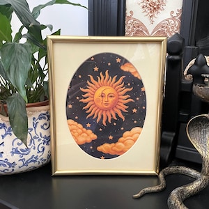 May include: A rectangular gold-toned frame containing an oval-shaped print of a stylized sun and moon face set against a dark blue starry night sky with orange clouds. The artwork features a central sun with a crescent moon profile overlay.