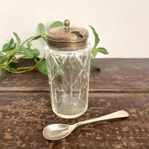 May include: A clear glass condiment jar with a diamond-cut pattern and a metallic lid featuring a small knob and a slot for a spoon. A matching metallic spoon with a smooth, rounded bowl and a slender handle is placed in front of the jar.