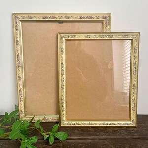 May include: Two rectangular picture frames with a gold finish and a decorative border featuring a repeating pattern of small pink roses and green leaves. The frames are positioned one behind the other, each containing a plain brown backing board.