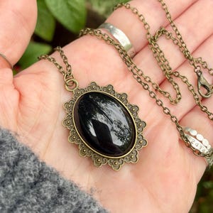 May include: An antique-style necklace with a large, oval black stone pendant. The pendant is set in a decorative, bronze-colored frame and hangs from a matching chain. The necklace is displayed on a hand.