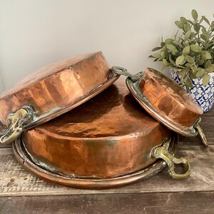 May include: Three antique copper pans with brass handles are stacked. The pans vary in size, with a rich copper colour and visible signs of age. The handles are ornate, adding to the vintage aesthetic. The pans are on a wooden surface.