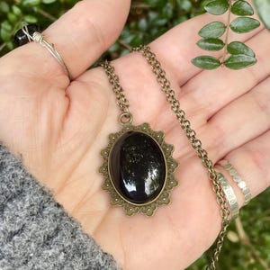 Pode incluir: Um colar de estilo vintage com um pingente de pedra oval escura. O pingente é fixado em uma moldura de filigrana de cor bronze e pendurado em uma corrente combinando. Anéis de prata também são visíveis.