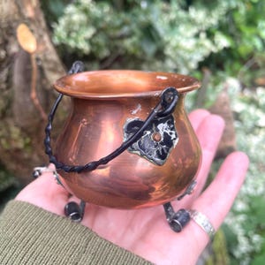 May include: A small, round copper cauldron featuring a dark, twisted metal handle and three short, sturdy metal legs. A decorative, heart-shaped black enamel patch with a small central stud is attached to the front of the polished copper body.