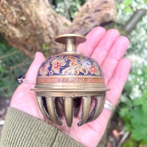 May include: A brass bell featuring a dome-shaped top with an intricate blue, red, and gold floral pattern. The bell has a flat circular handle at the apex and a series of vertical, curved metal slats forming the lower section of the bell structure.