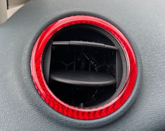 May include: Red carbon fiber trim ring for a car air vent. The ring is made of a durable material and has a sleek, modern design.