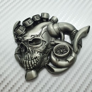 May include: A silver metal skull with a turbocharger and exhaust pipes. The skull has a detailed design and is a popular automotive accessory.