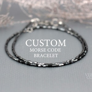 Puede incluir: Un brazalete de cuentas plateadas y negras con el texto "CUSTOM MORSE CODE BRACELET" sobre un fondo gris.