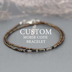 Puede incluir: Un delicado brazalete de doble cadena hecho con pequeñas cuentas iridiscentes en tonos marrones, negros y plateados. El brazalete está etiquetado como "CUSTOM MORSE CODE BRACELET" y "VERY CODE".