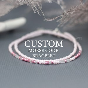 May include: A custom Morse code bracelet made with pink and white beads. The bracelet is on a gray surface with the text "CUSTOM MORSE CODE BRACELET" above it.