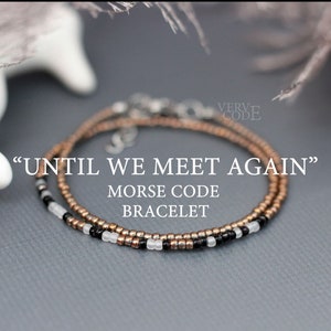 Puede incluir: Un brazalete de código Morse con el mensaje "UNTIL WE MEET AGAIN". El brazalete presenta pequeñas cuentas metálicas en oro, negro y blanco. La joya se exhibe sobre una superficie gris, con elementos borrosos en el fondo.