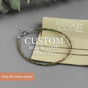 Puede incluir: Un brazalete de código Morse personalizado hecho con pequeñas cuentas verdes, marrones y doradas. El brazalete está sobre una bolsa de tela beige con el texto "VERVE CODE" impreso en ella. El texto "CUSTOM MORSE CODE BRACELET" está impreso debajo del brazalete. El texto "haz clic para ver más colores" está impreso debajo de la bolsa.