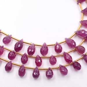 May include: Three strands of faceted, teardrop-shaped, deep pink gemstones strung on gold-colored wire. The gemstones are arranged in a layered pattern against a white background, showcasing their polished surfaces.