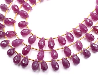 May include: Close-up of three strands of faceted, teardrop-shaped ruby beads. The beads are a deep magenta colour and strung on thin gold-coloured wire. The beads are arranged in a curved pattern against a white background.