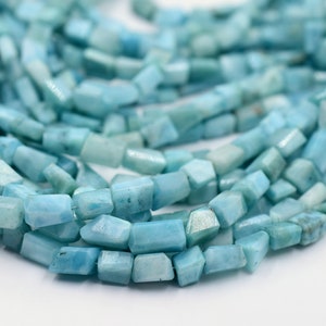 Könnte beinhalten: Nahaufnahme eines Strangs hellblauer Edelsteinperlen. Die Perlen sind rechteckig mit facettierten Kanten und sind aufgefädelt. Die Perlen haben ein leicht durchscheinendes Aussehen und variieren in den Farbtönen Blau und Aqua.