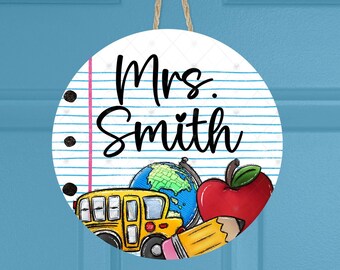 May include: A round, white door hanger with the text "Mrs. Smith" written in black cursive. The background is a pink and white striped pattern that resembles notebook paper. There are black dots and a yellow school bus, a red apple, a pencil, and a globe on the hanger.