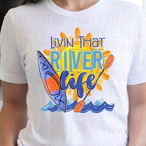 Könnte beinhalten: Weißes T-Shirt mit einem Aquarell-Design eines Kajaks, einer Sonne und Wellen. Der Text "Livin' That River Life" ist in einer bunten Schrift geschrieben.