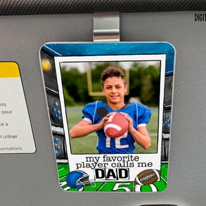 May include: A rectangular car visor clip featuring a photo of a young football player in a blue uniform holding a football. The clip has a football field design and the text "my favorite player calls me DAD".