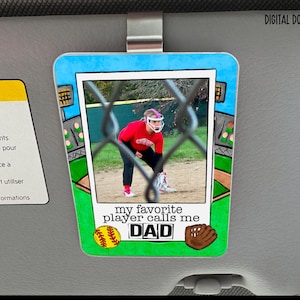 May include: A rectangular car visor clip featuring a softball player in a red uniform behind a chain-link fence. The clip has a baseball field design with a baseball, glove, and the text "my favourite player calls me DAD."