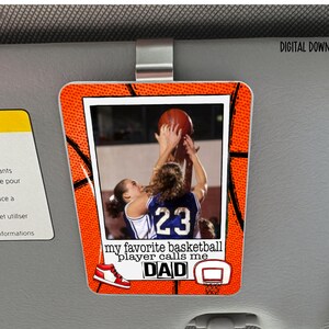 Peut inclure: Un pare-soleil de voiture rectangulaire avec un motif de basket-ball et une photo de deux joueurs de basket-ball. Le texte indique "my favorite basketball player calls me DAD". Le pare-soleil est fixé à un pare-soleil de voiture.
