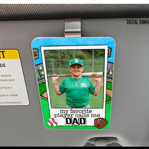 Op de afbeelding: Een auto zonneklep clip met een honkbal thema, met een foto van een jonge honkbalspeler met een knuppel. De tekst "my favorite player calls me DAD" wordt weergegeven. De clip heeft honkbal- en handschoenafbeeldingen.