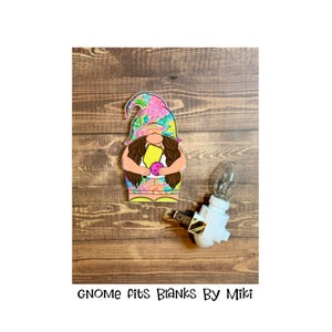 May include: A colorful floral gnome figurine holding a yellow and pink object. The gnome is sitting on a wooden surface next to a white nightlight with a gold accent.