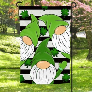 May include: A garden flag with a black and white striped background and three gnomes wearing green hats and white beards. The gnomes are surrounded by green shamrocks.