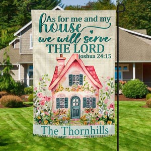 Op de afbeelding: Een tuinvaandel met een aquarel illustratie van een roze huis met een rood dak, omringd door bloemen. De vlag heeft de tekst "As for me and my house we will serve the Lord Joshua 24:15" en "The Thornhills".